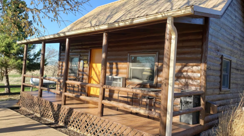 A rustic log cabin with a wooden porch, featuring chairs and an orange door, set against a clear blue sky.